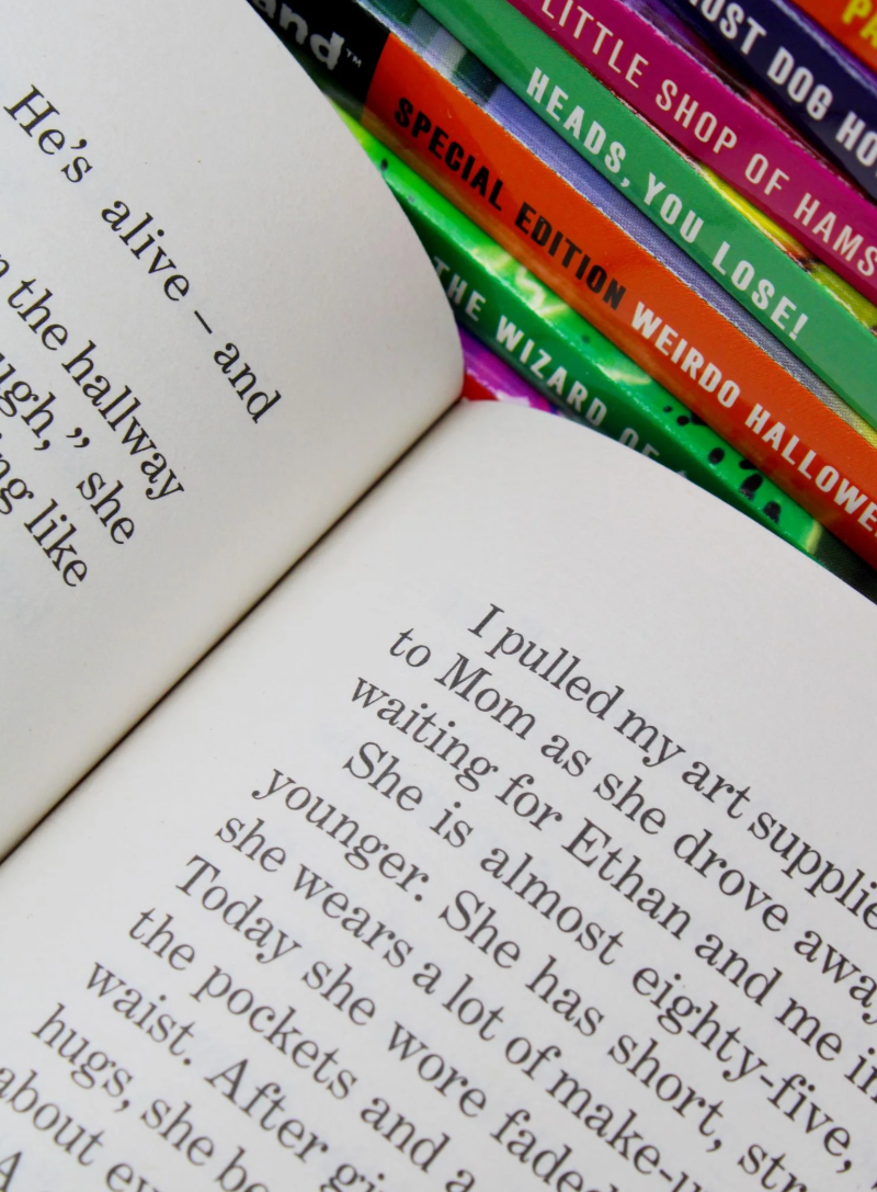 An open book showing printed text on the pages, with a row of colorful hardcover books stacked along the top right edge against a white background.
