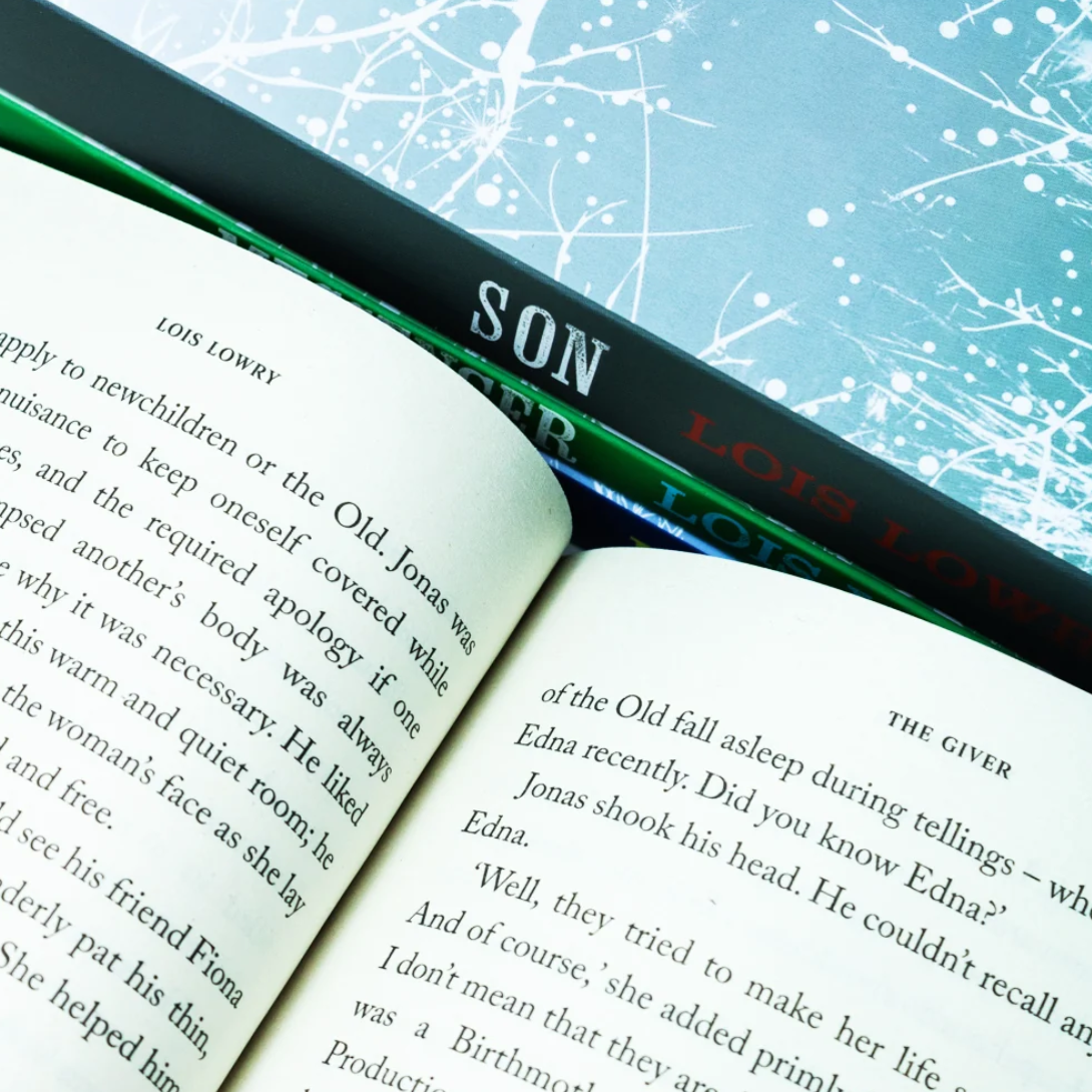 An open book with printed text on cream pages lies diagonally across a turquoise surface, partially covering a black book with green and white lettering that reads 'SON' and 'LOIS LOWRY.'.